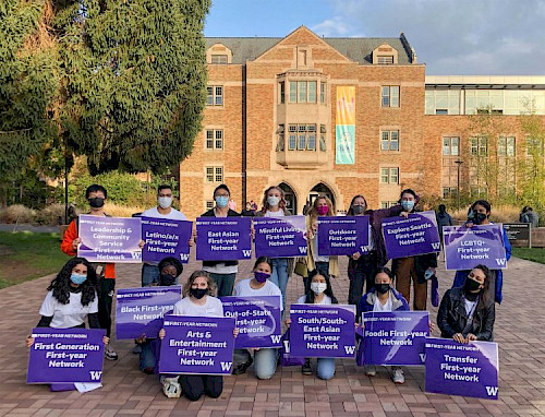 Fourteen students standing together, holding purple signs with various first year network groups written on them
