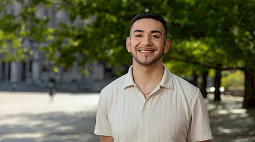 picture of person with tan shirt in front a green trees