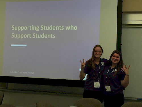Two students holding up the "W" hand sign in front of a projection reading "supporting students who support students"